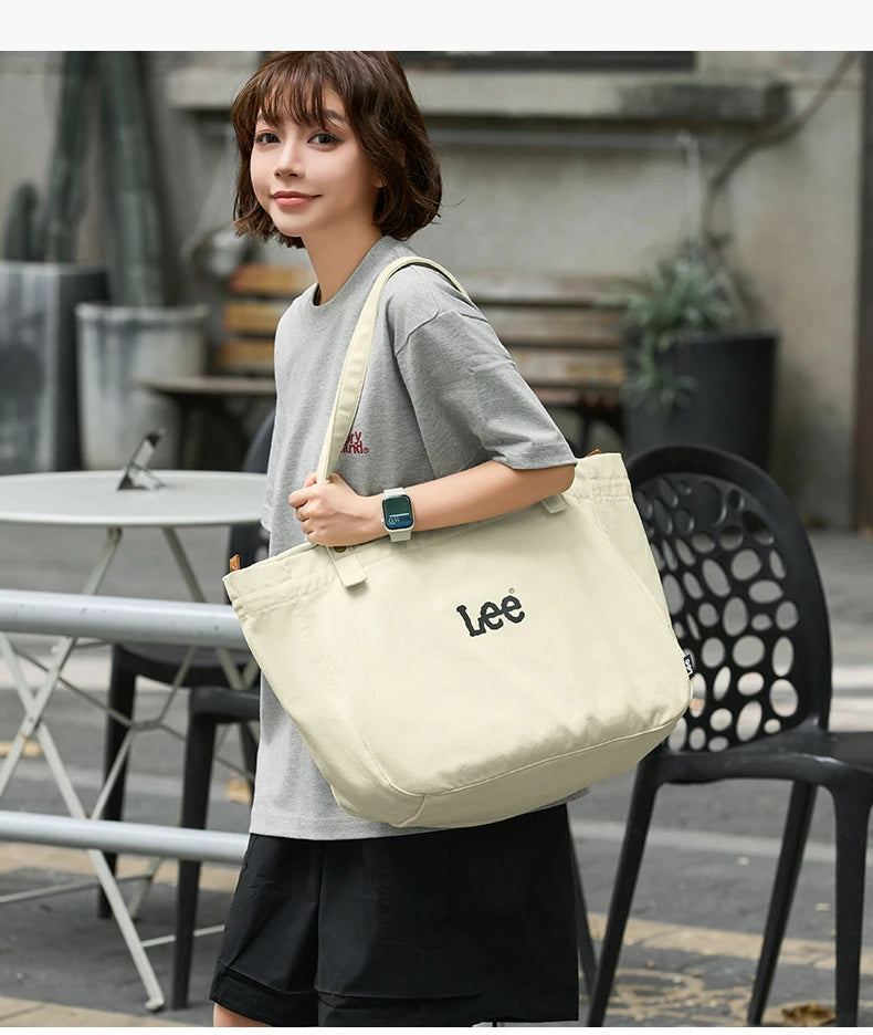 A woman wearing a grey t-shirt and black shorts holds a cream colored Lee Oxford cloth tote bag.