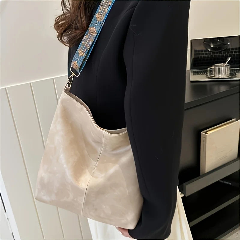 A beige leather shoulder bag with a blue embroidered strap is shown hanging from a black blazer sleeve.