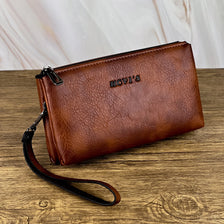 Portefeuille pochette en cuir marron avec fermeture éclair et sangle posé sur une surface en bois.