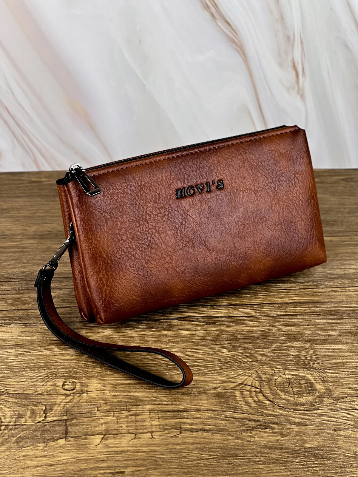 Portefeuille pochette en cuir marron avec fermeture éclair et sangle posé sur une surface en bois.