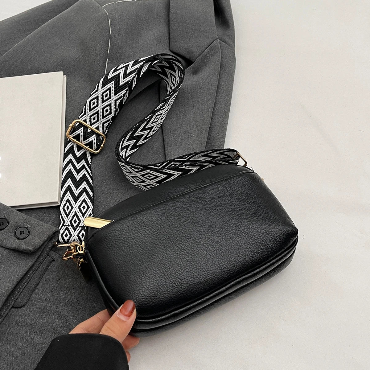 A hand holds a black vegan PU crossbody bag with a patterned strap and a gray blazer.