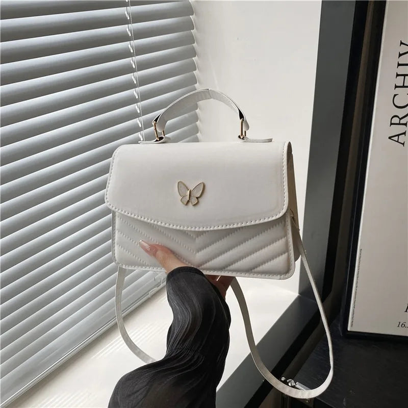 A white quilted shoulder messenger bag with a silver hardware is held by a person in front of a window blind.