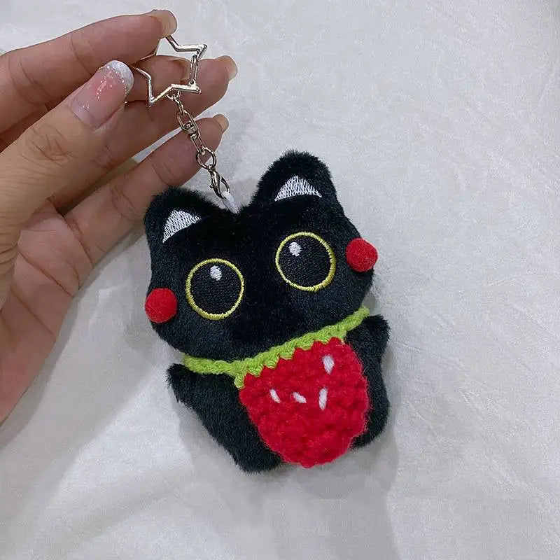 Colgante de gato negro de peluche con detalles rojos y cadena plateada sostenido en una mano.