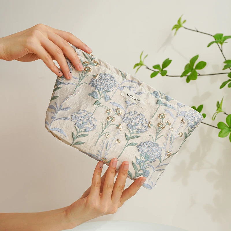 A hand is holding a light blue floral canvas makeup bag against a white background with green foliage.