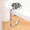 A silver zinc alloy folding handbag hook with a black and white floral pattern is displayed on a white surface.