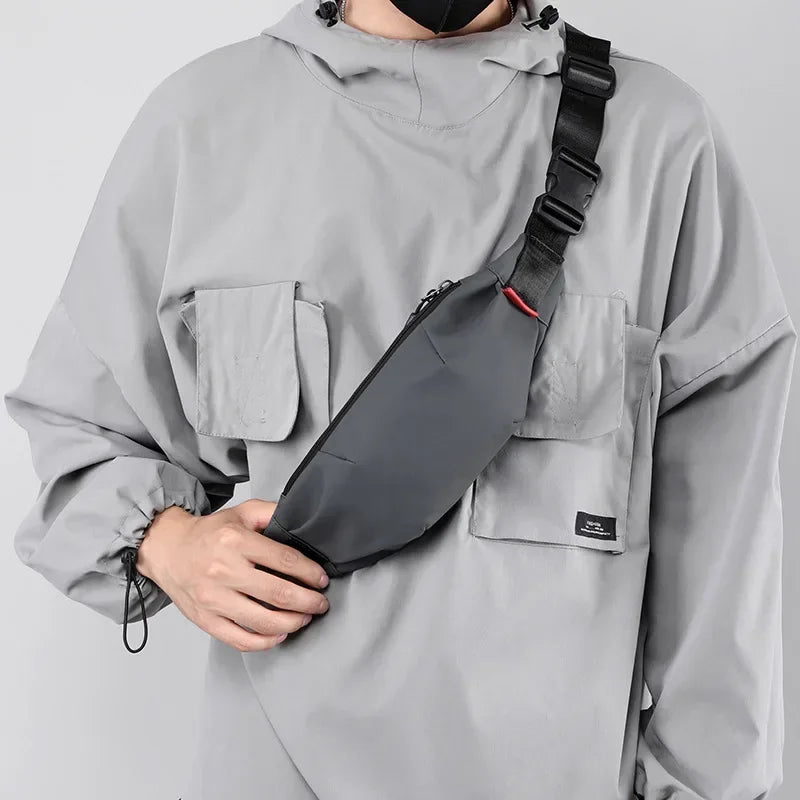 A person wearing a gray hoodie is holding a black water-resistant nylon waist bag with an adjustable strap.