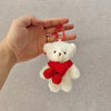 A white plush bear with a red scarf is being held by a hand, attached to a keychain.