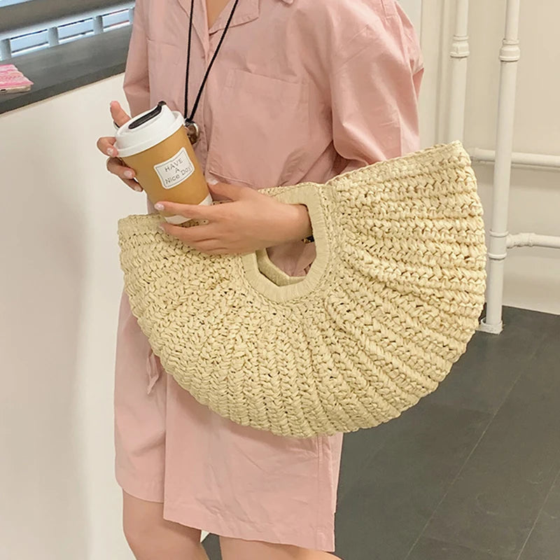 A person holds a beige woven rattan half-moon beach handbag against a light pink dress.