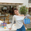 A young woman wearing a white off-the-shoulder top and jeans stands in front of a glass storefront with greenery.