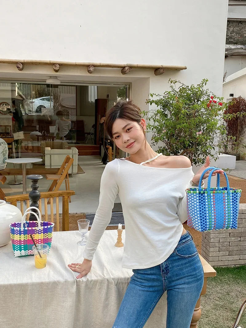 A young woman wearing a white off-the-shoulder top and jeans stands in front of a glass storefront with greenery.