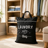 A black laundry bag with a handle is displayed in front of wooden shelving and clothing racks.