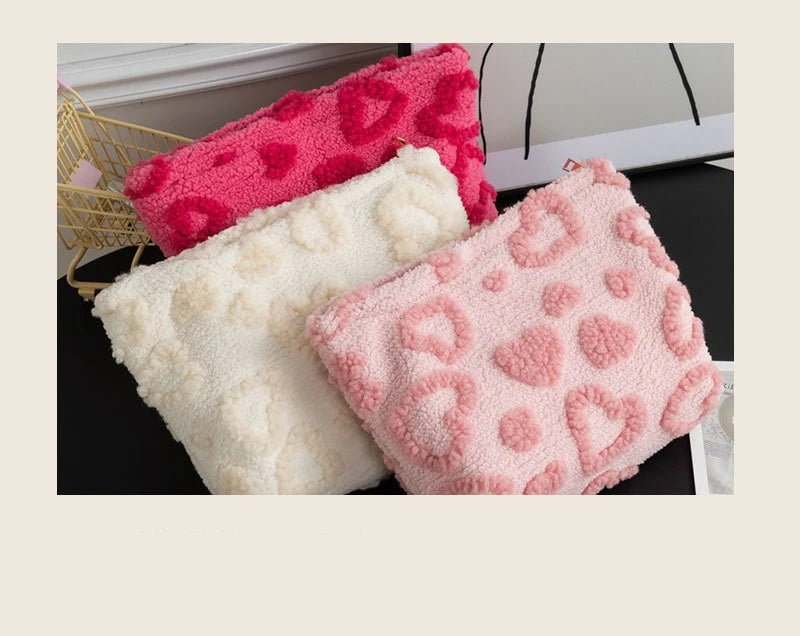 Three plush heart-pattern storage bags in pink, white, and bright pink are displayed on a dark surface.