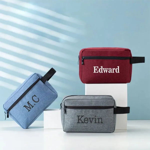 Three personalized embroidered travel cosmetic bags in navy, maroon, and gray resting on a white surface.