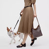 A woman wearing a beige skirt and black boots holds a brown geometric embroidery hobo shoulder bag while a small dog stands b