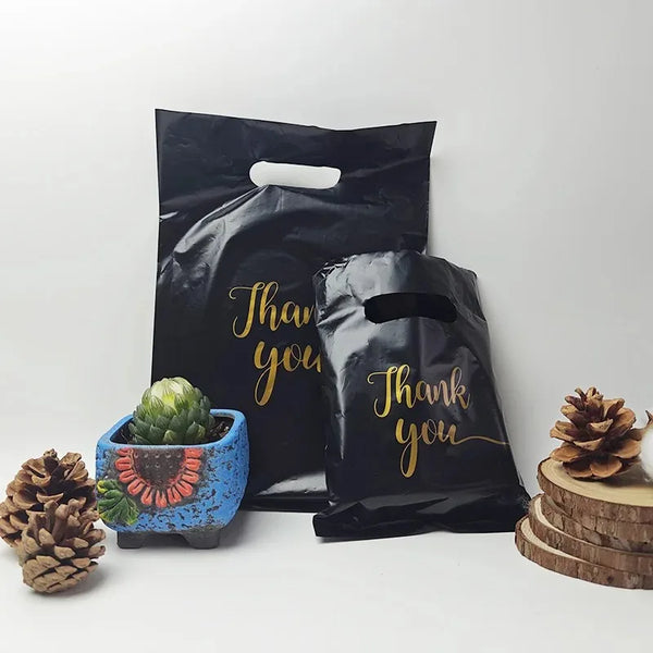 Black bulk thank you gift bags with gold lettering next to a succulent and pine cone