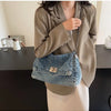 A woman wearing a beige blazer and light pants holds a denim tote bag with a silver chain strap.