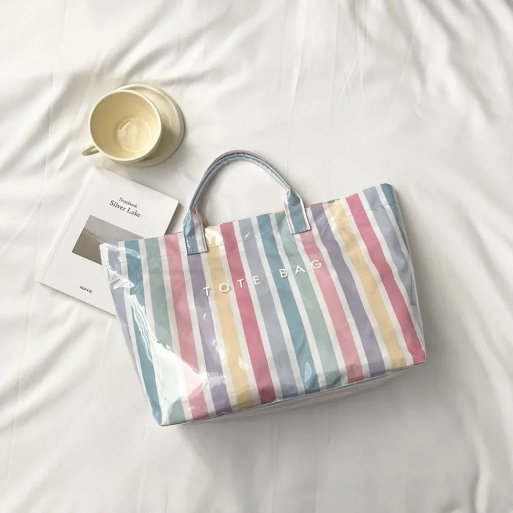 A striped transparent PVC tote bag rests on a white sheet next to a cup.