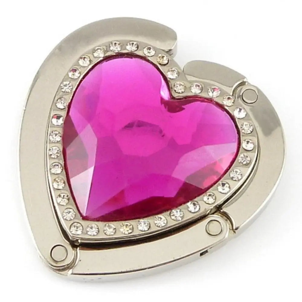 A heart-shaped metal fold-over bag hook with a faceted magenta gemstone set in a silver frame.