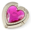 A heart-shaped metal fold-over bag hook with a faceted magenta gemstone set in a silver frame.