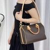 A woman holds a brown leather shoulder bag with a tan strap against a white curtain backdrop.