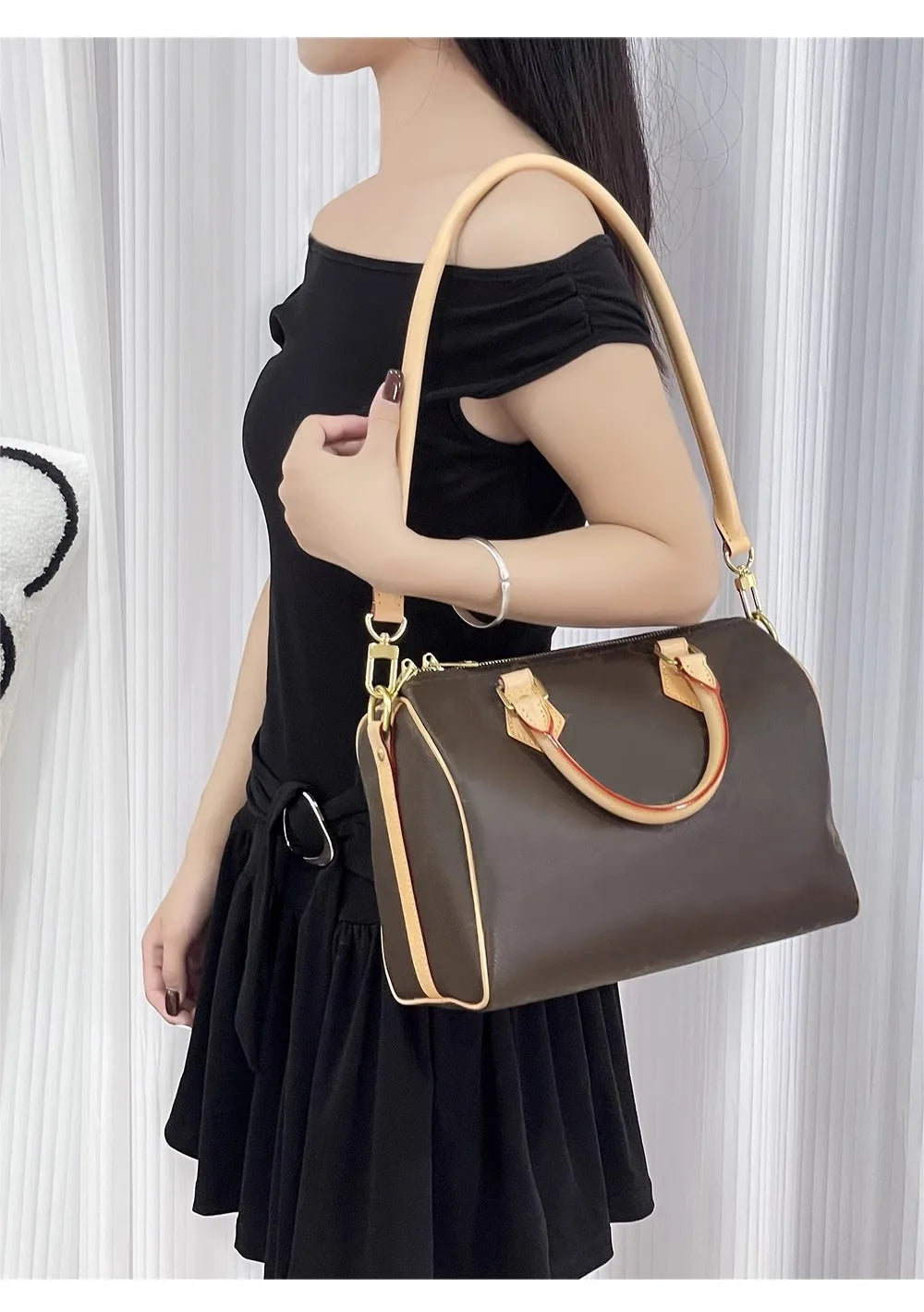 A woman holds a brown leather shoulder bag with a tan strap against a white curtain backdrop.