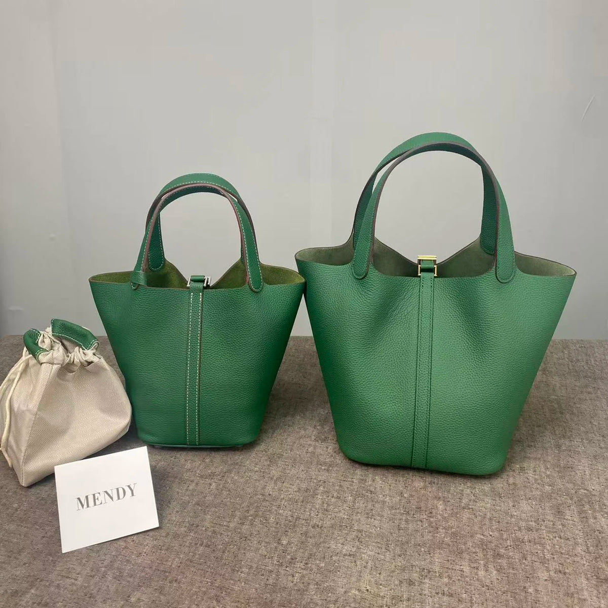A pair of dark green genuine leather bucket bags are displayed on a gray surface next to a small white card labeled MENDY.