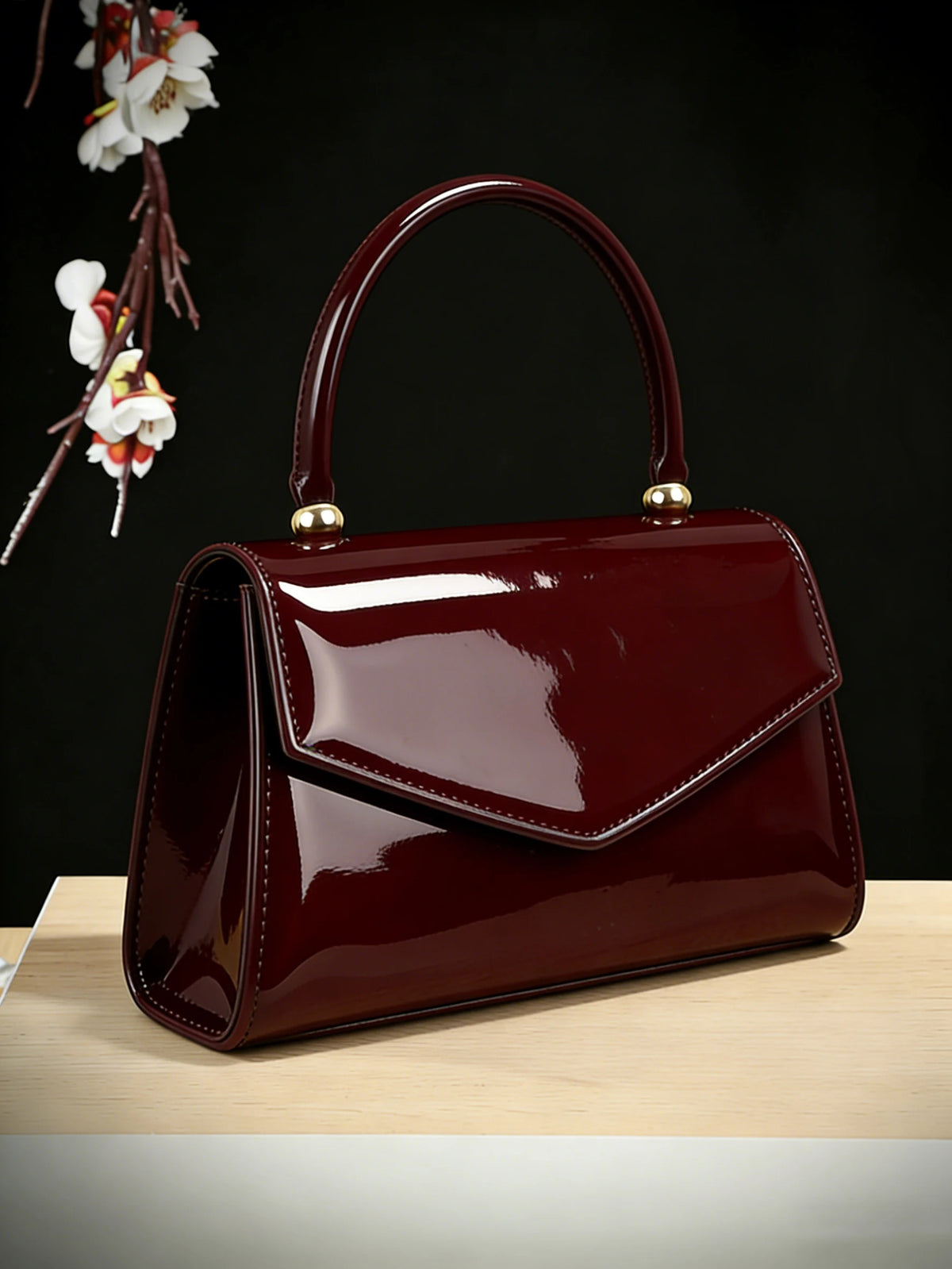 A glossy, deep burgundy evening handbag with a leather texture is displayed on a light wooden surface.
