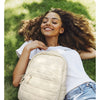 A woman is smiling while holding a beige quilted casual backpack outdoors on green grass.