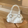 A white PU leather shoulder bag with silver rivets resting on a light wooden surface.