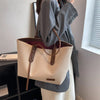 A person holds a large beige canvas shoulder tote bag with brown leather straps next to a window.