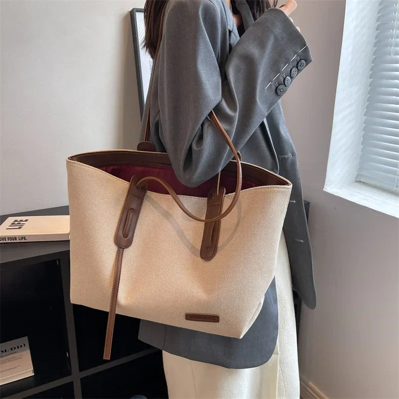 A person holds a large beige canvas shoulder tote bag with brown leather straps next to a window.