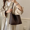 A woman wearing a beige trench coat holds a dark brown PU bucket shoulder bag.