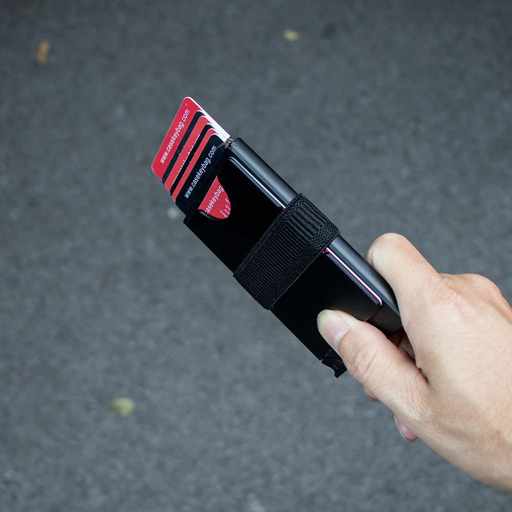 A hand holds an RFID blocking metal pop-up card wallet with a red and black design against a dark background.