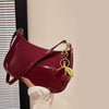 A deep burgundy PU leather shoulder bag with a gold cherry pendant is held by a hand against a light wall.