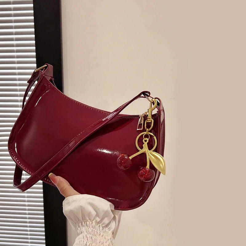 A deep burgundy PU leather shoulder bag with a gold cherry pendant is held by a hand against a light wall.