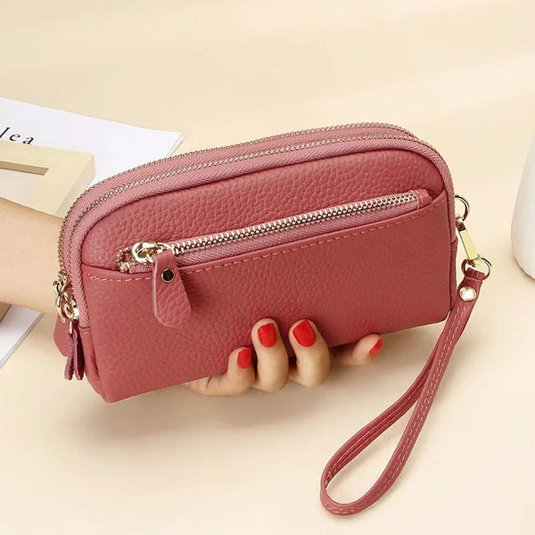 A pink genuine cowhide leather double-zip wristlet wallet with a brown leather strap is held by a hand.