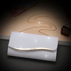 A gold chain and a thin gold bar are displayed on a light gray envelope clutch with a dark glass bottle in the background.
