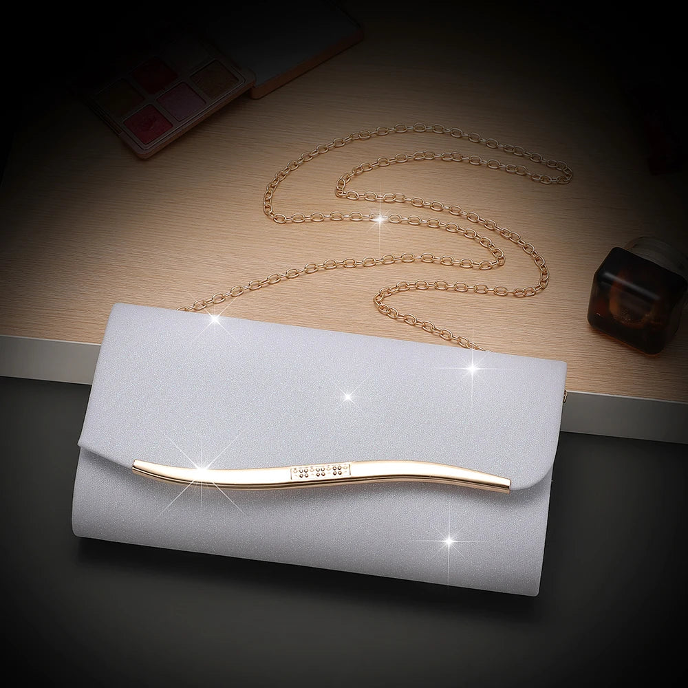 A gold chain and a thin gold bar are displayed on a light gray envelope clutch with a dark glass bottle in the background.