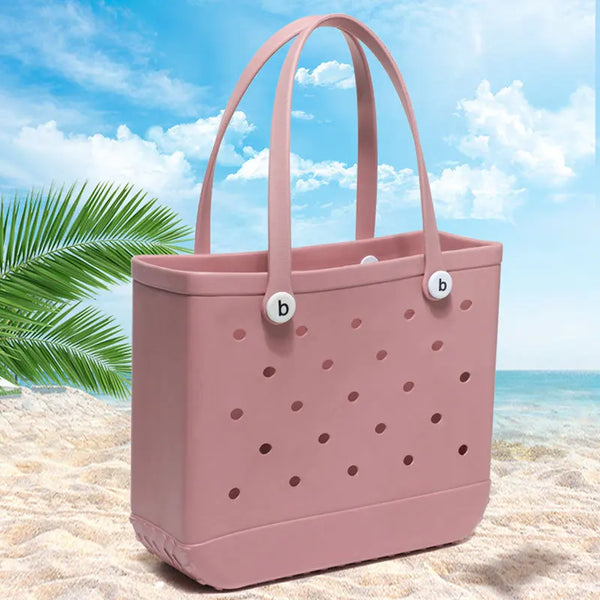 A pink portable EVA beach tote bag with perforated details resting on light sand near the ocean.