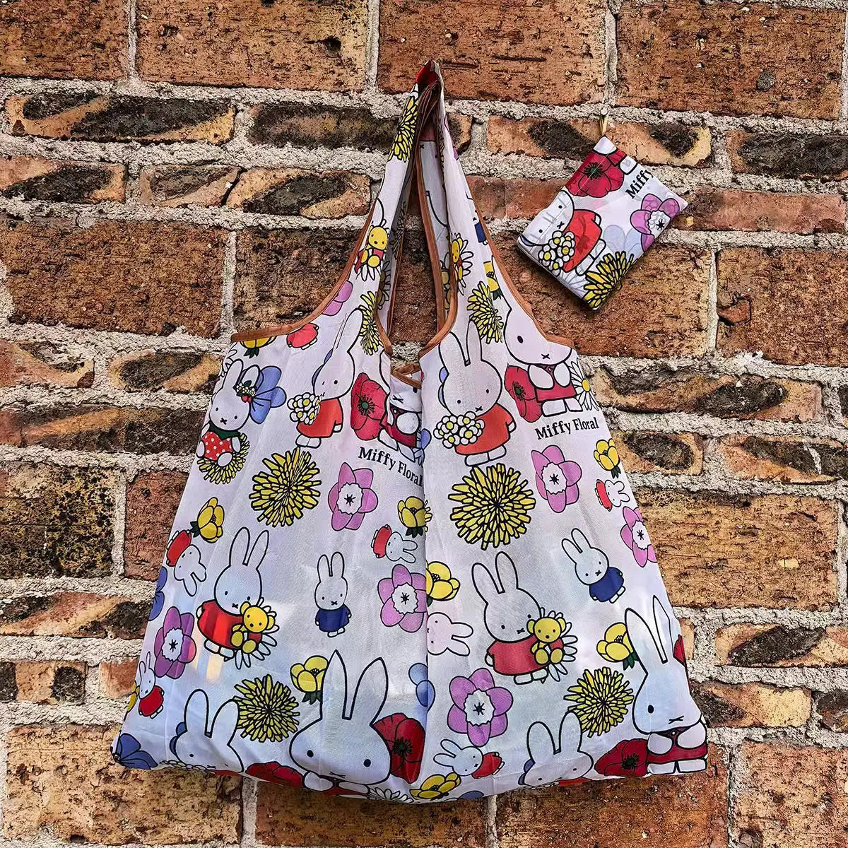 A white Miffy folding shopping bag featuring colorful illustrations of Miffy and flowers, displayed against a brick wall.