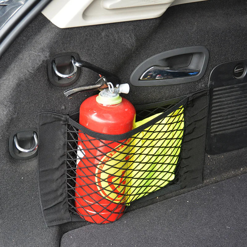 A red bottle is stored in a black mesh bag inside a car trunk organizer net.
