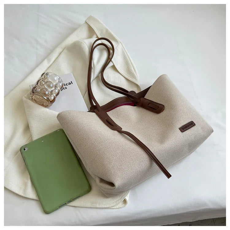 A beige canvas shoulder tote bag with brown leather straps and a green tablet resting on the fabric.