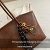 A brown leather tote bag with a gold-toned hanging ornament and strap resting on a white surface.
