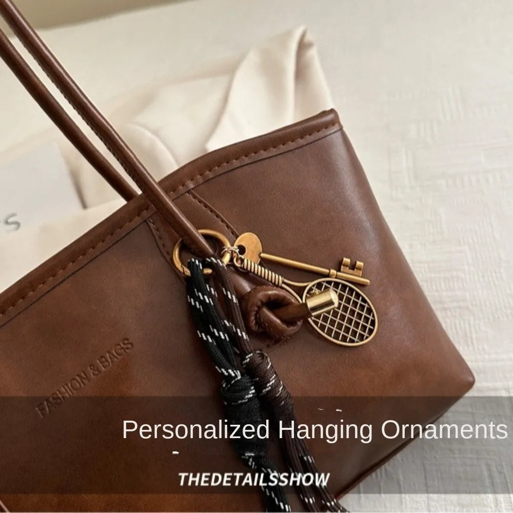 A brown leather tote bag with a gold-toned hanging ornament and strap resting on a white surface.