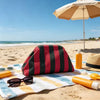 An elegant knit cloud makeup bag rests on a striped towel with sunglasses and sunscreen on a sandy beach.