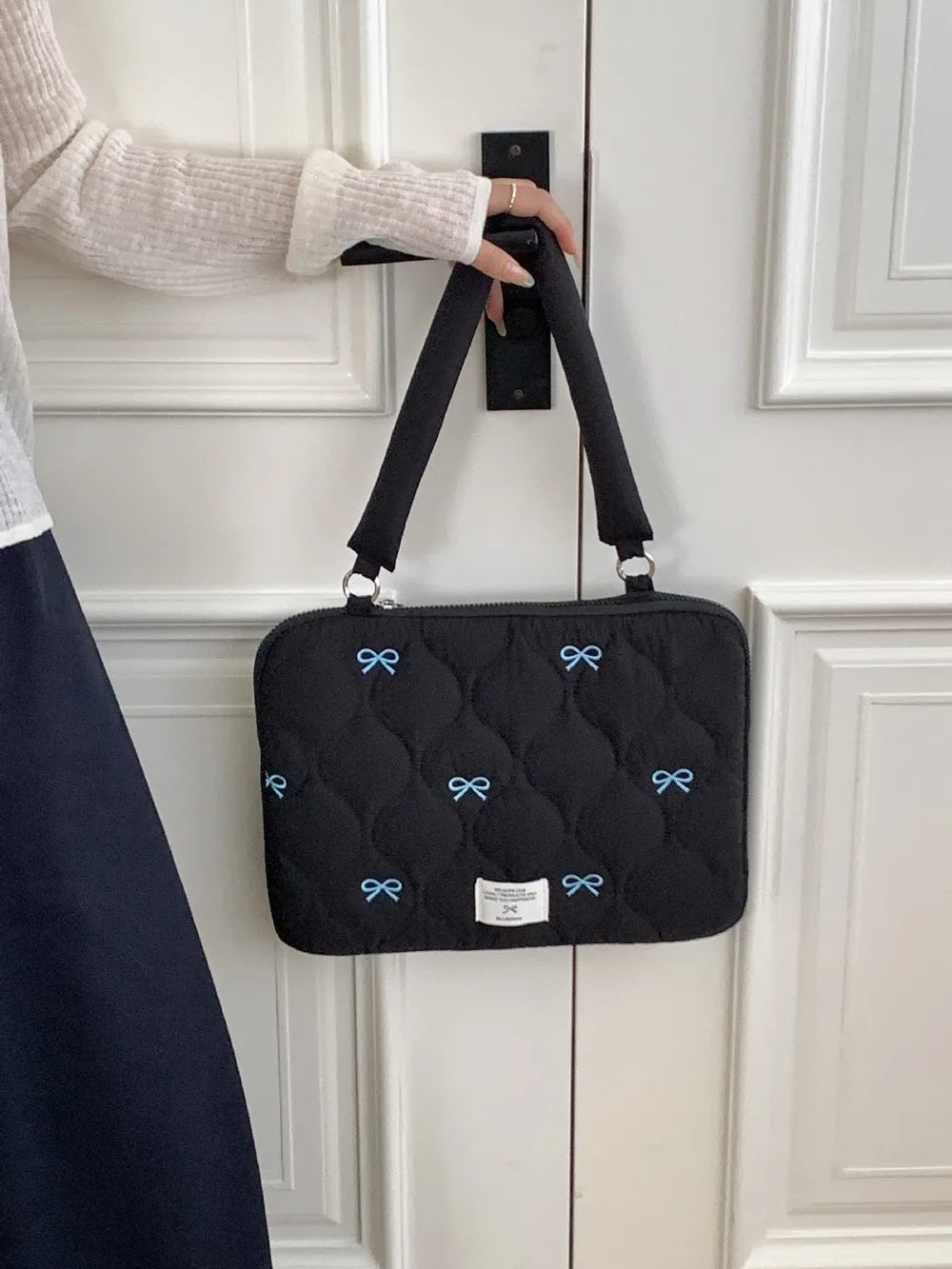 Bolso para portátil acolchado negro con detalles de lazo azul colgado en un marco de puerta blanca.