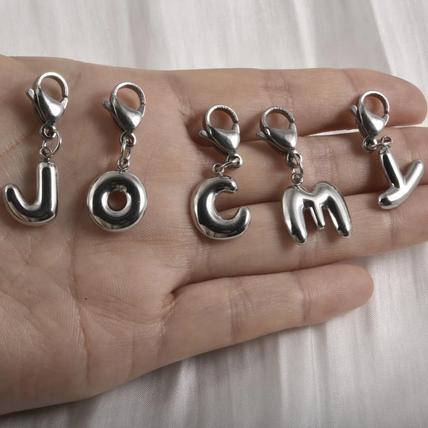 A hand holds several stainless steel alphabet letter charms with lobster clasps.