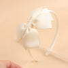 A close-up of a cream colored fabric bow headband with a thin elastic band on a light background.