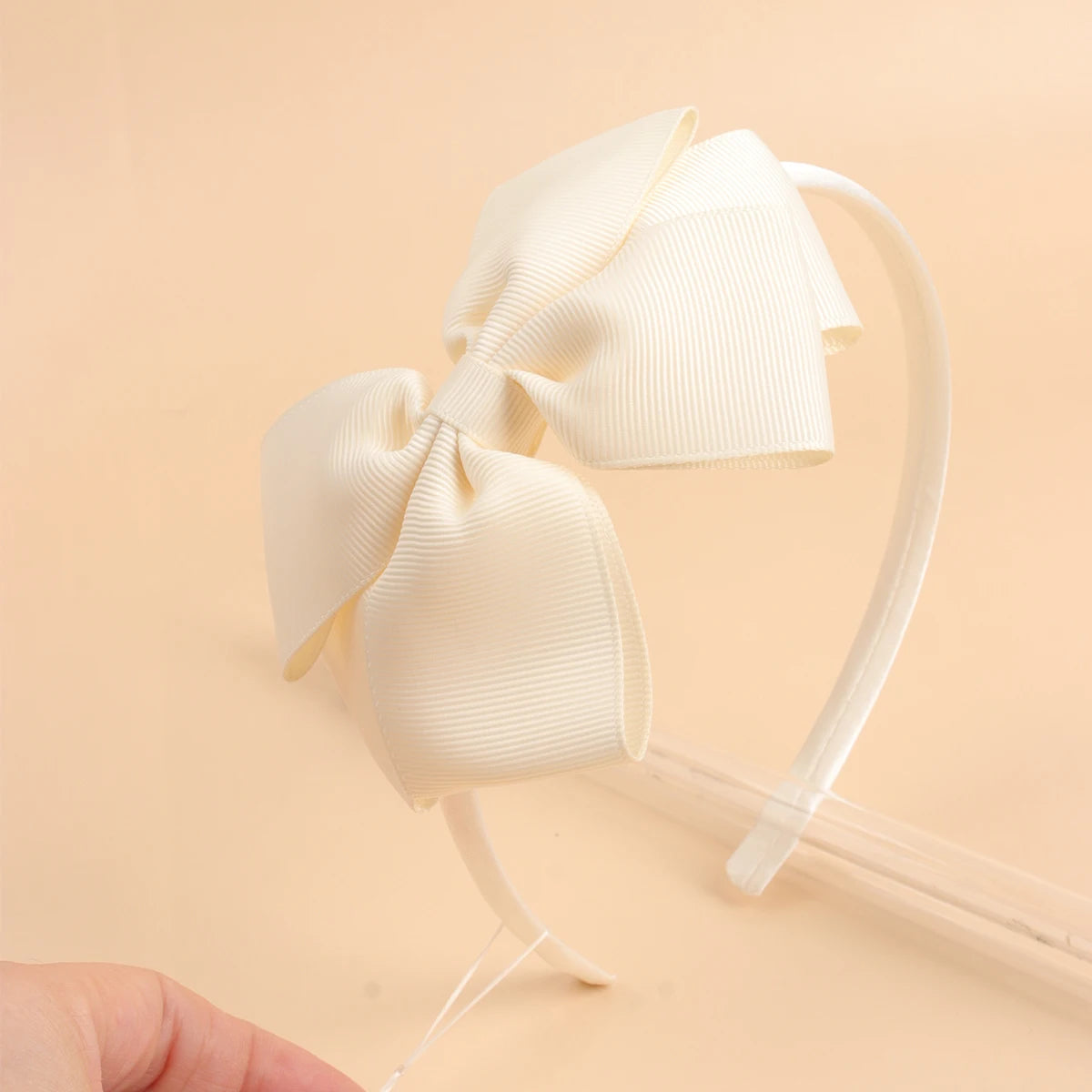 A close-up of a cream colored fabric bow headband with a thin elastic band on a light background.