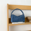 A deep blue faux suede crossbody bag resting on a wooden shelf with a stack of books.